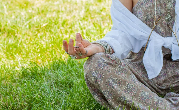Woman is meditating sitting in Lotus pose on grass at the park. Boho style with accessories, rings and bracelets. — Stok fotoğraf