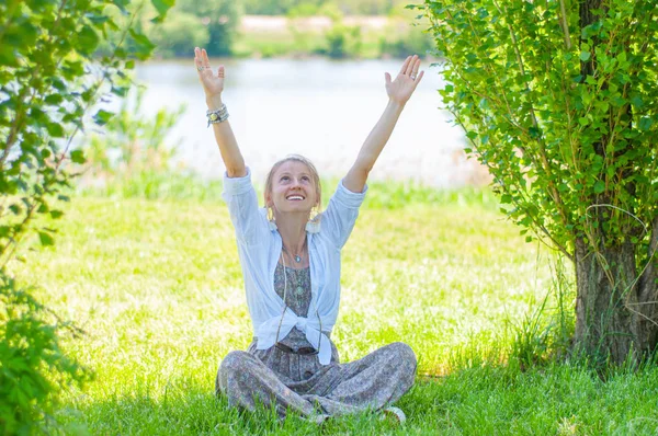 Femeia frumoasă își ridică mâinile este așezată pe iarbă se bucură de o zi însorită în parc. Fata sta în Lotus pozează în aer liber . — Fotografie, imagine de stoc