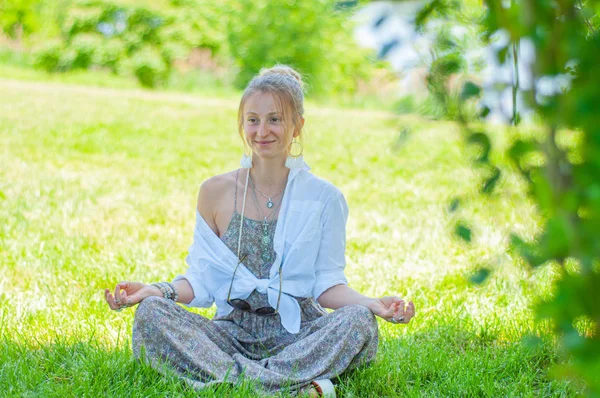 Femeia fericită meditează stând în Lotus pozând pe iarbă. Frumoasă femeie în stil boho cu accesorii care se bucură de o zi însorită în parc . — Fotografie, imagine de stoc