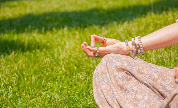 Mâna feminină cu inele și brățări accesorii stil boho. Femeia meditează stând în Lotus pozând pe iarbă . — Fotografie, imagine de stoc