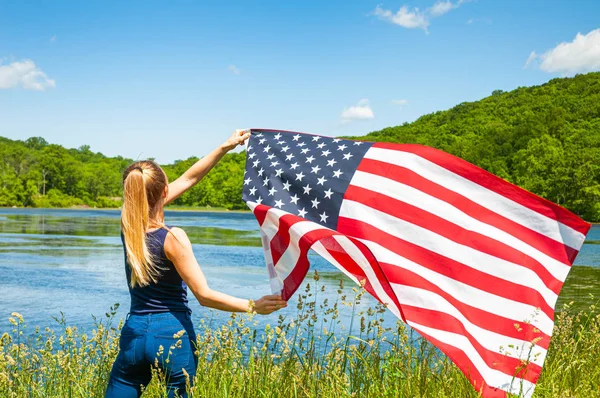 4 lipca. Młoda kobieta trzyma amerykańską flagę na tle jeziora. — Zdjęcie stockowe