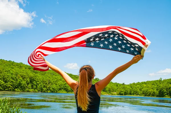 4 lipca. Młoda kobieta trzyma amerykańską flagę na tle jeziora. — Zdjęcie stockowe