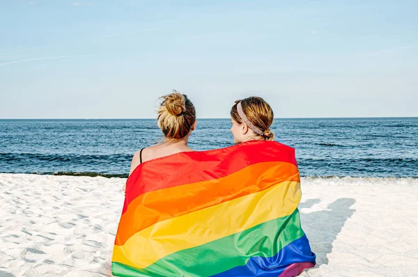 Pareja de lesbianas enamoradas en la playa con una bandera de arco iris, símbolo de la comunidad LGBT — Foto de Stock