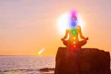 Kadın gün batımında taş üzerinde parlayan yedi çakra ile meditasyon olduğunu. Kadının silueti sahilde yoga yapıyor.