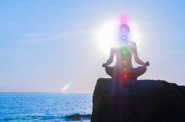 Kadın gün batımında taş üzerinde parlayan yedi çakra ile meditasyon olduğunu. Kadının silueti sahilde yoga yapıyor.