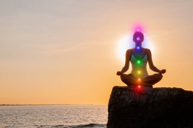 Kadın gün batımında taş üzerinde parlayan yedi çakra ile meditasyon olduğunu. Kadının silueti sahilde yoga yapıyor.