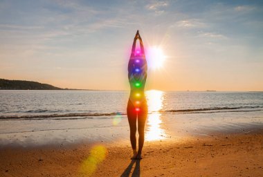 Kadın sahilde parlayan yedi çakra ile meditasyon yapıyor. Kadının silueti gün batımında yoga yapıyor.