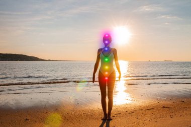 Kadın sahilde parlayan yedi çakra ile meditasyon yapıyor. Kadının silueti gün batımında yoga yapıyor.