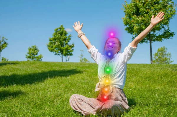 Mujer feliz está meditando en la pose de loto con siete chakras brillantes en la hierba. La mujer está practicando yoga en el parque . — Foto de Stock