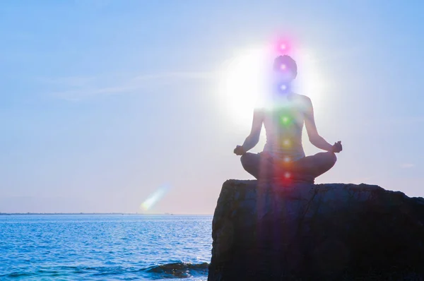 Vrouw is mediteren met gloeiende zeven chakra's op steen bij zonsondergang. Silhouet van de vrouw is het beoefenen van yoga op het strand. — Stockfoto