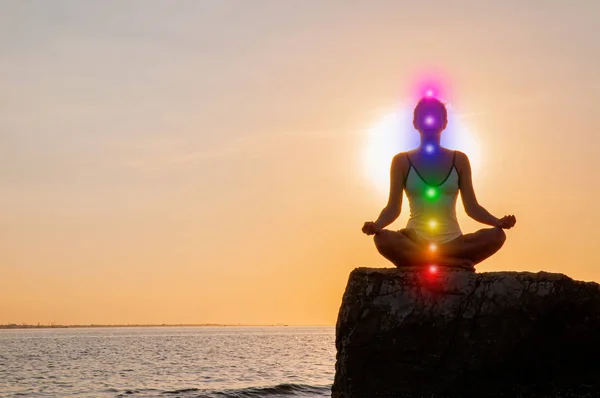 Vrouw is mediteren met gloeiende zeven chakra's op steen bij zonsondergang. Silhouet van de vrouw is het beoefenen van yoga op het strand. — Stockfoto