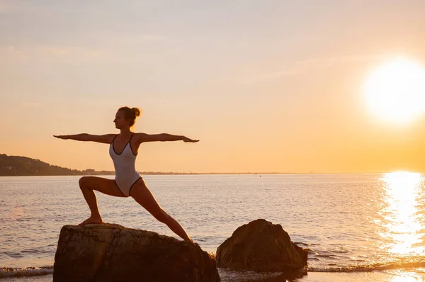 Frau praktiziert Yoga bei Sonnenuntergang auf Stein. Silhouette einer Yoga-Frau am Strand bei Sonnenuntergang. — Stockfoto