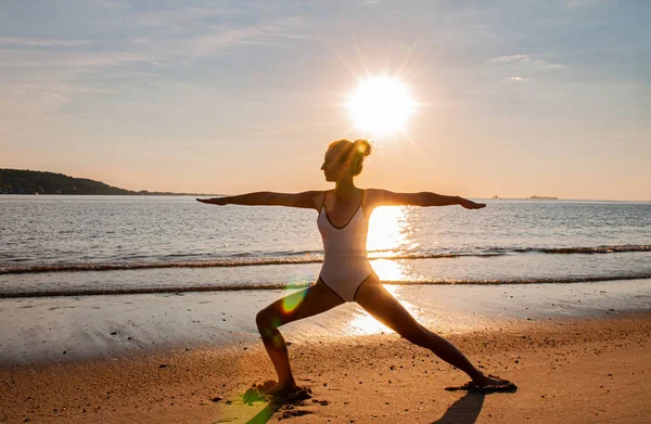 Silueta unei femei de yoga pe plajă la răsăritul soarelui. Femeia practică yoga la apusul soarelui pe malul mării . — Fotografie, imagine de stoc