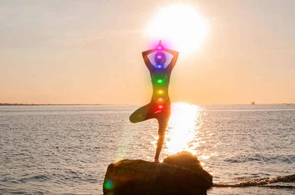 Kadın sahilde parlayan yedi çakra ile meditasyon yapıyor. Kadının silueti gün batımında yoga yapıyor.