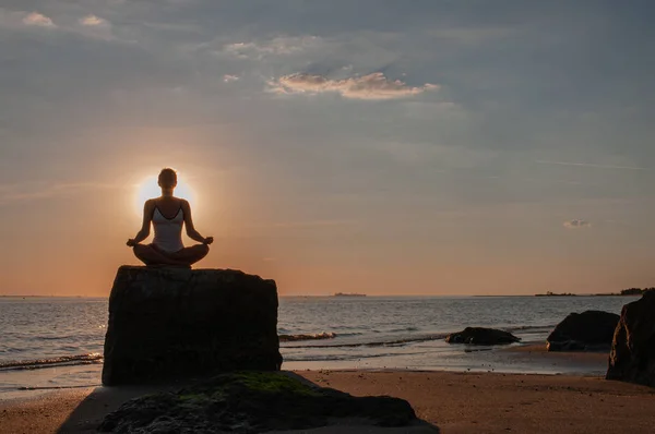 Femeia practică yoga așezată pe piatră în Lotus pozează la apusul soarelui. Silueta unei femei care meditează pe plajă — Fotografie, imagine de stoc