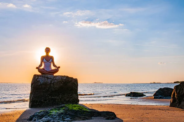 Kvinnan tränar yoga sitter på sten i Lotus pose vid solnedgången. Siluetten av kvinnan mediterar på stranden — Stockfoto