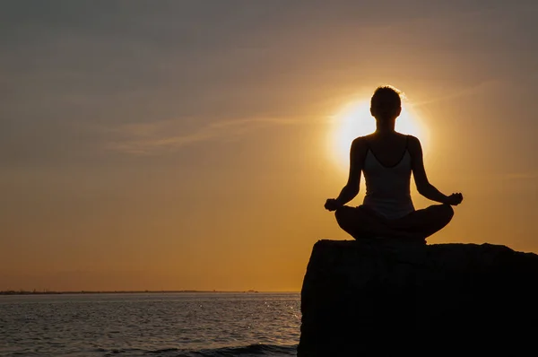 Donna sta praticando yoga seduto sulla pietra in posa Lotus al tramonto. Silhouette di donna che medita sulla spiaggia — Foto Stock