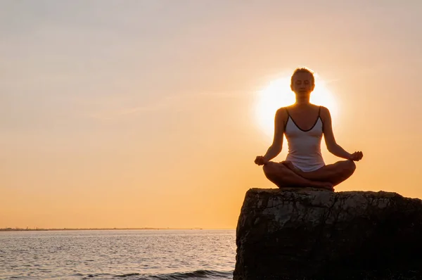 Mulher está praticando ioga sentado em pedra em pose de lótus ao pôr do sol. Silhueta de mulher meditando na praia — Fotografia de Stock