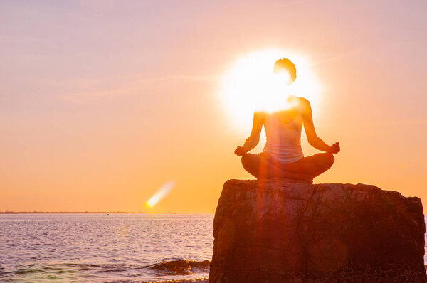 Женщина практикует йогу сидя на камне в позе Лотоса на закате. Силуэт женщины медитирующей на пляже
 
