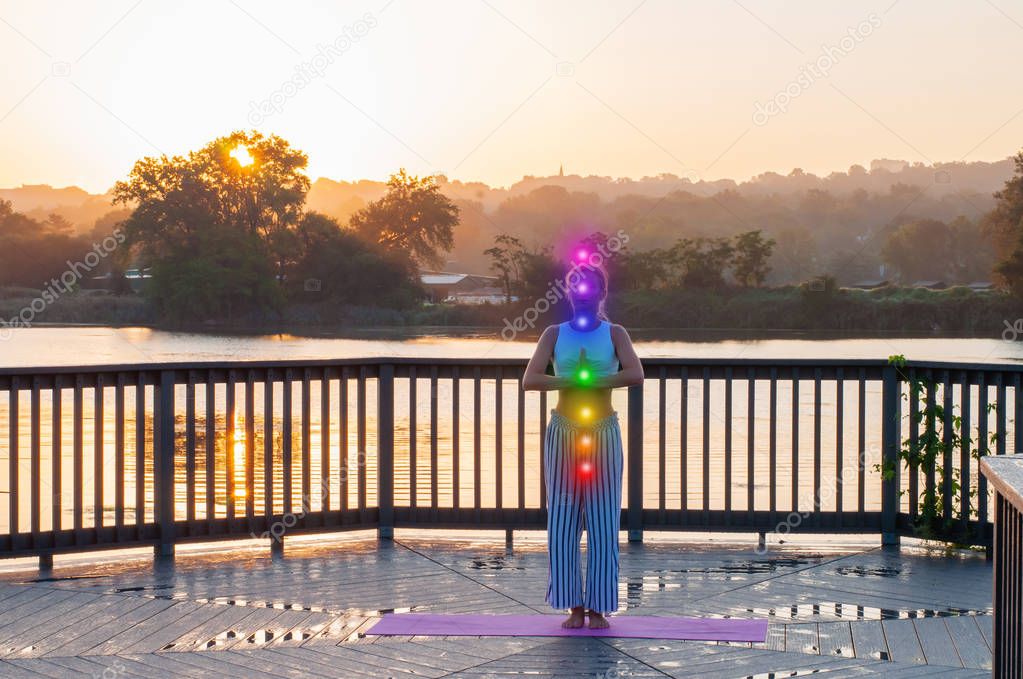 Woman is meditating with glowing seven chakras.  Beautiful  woman is practicing yoga on the lake.
