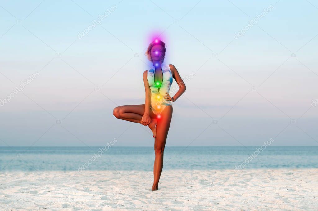 Woman is meditating with glowing seven chakras on the beach. Beautiful woman is practicing yoga at sunset.