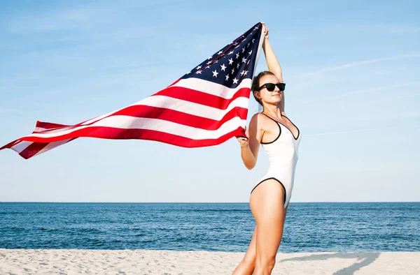 Bela mulher patriótica segurando uma bandeira americana na praia. Dia da independência dos EUA, 4 de julho . — Fotografia de Stock