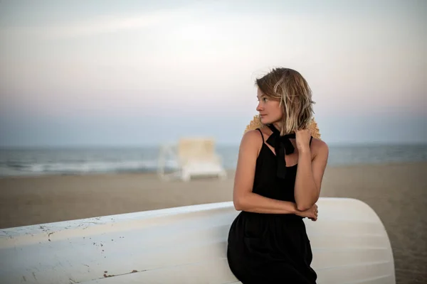 Beautiful woman in black summer dress on the beach near white wooden boat. — Stock Photo, Image