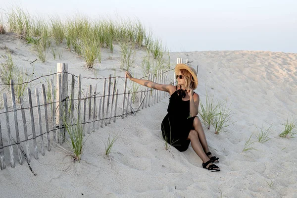 Beautiful woman in black summer dress and hat on the beach near wooden fence. — Stock Photo, Image