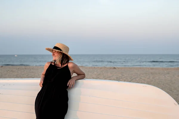 Beautiful woman in black summer dress and hat on the beach near wooden boat. — Stock Photo, Image