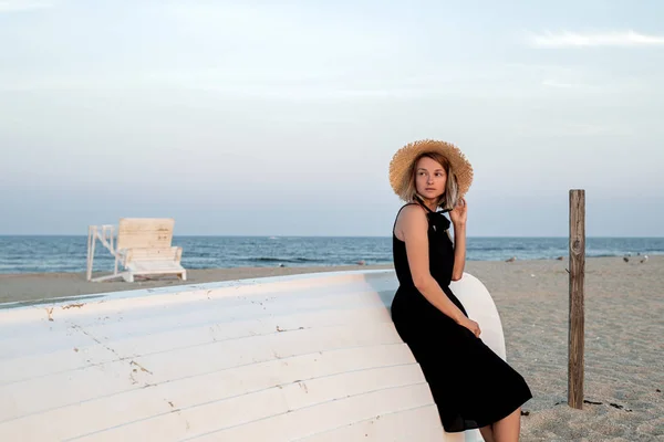 Beautiful woman in black summer dress and hat on the beach near wooden boat. — Stock Photo, Image