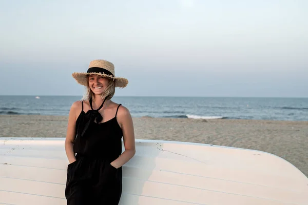 Beautiful woman in black summer dress and hat on the beach near wooden boat. — Stock Photo, Image