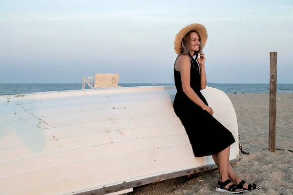 Beautiful woman in black summer dress and hat on the beach near wooden boat. — Stock Photo, Image