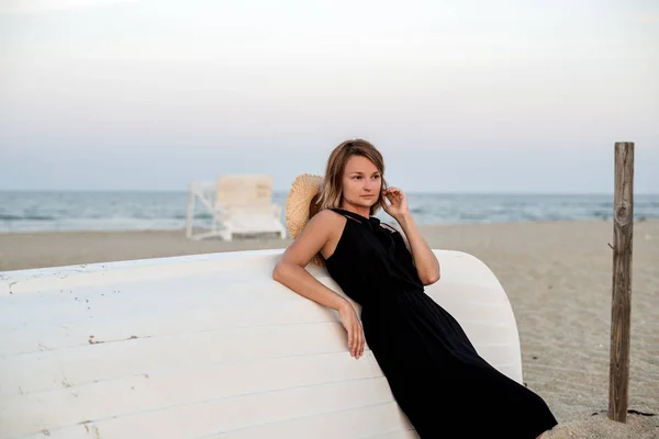 Beautiful woman in black summer dress on the beach near white wooden boat. — Stock Photo, Image