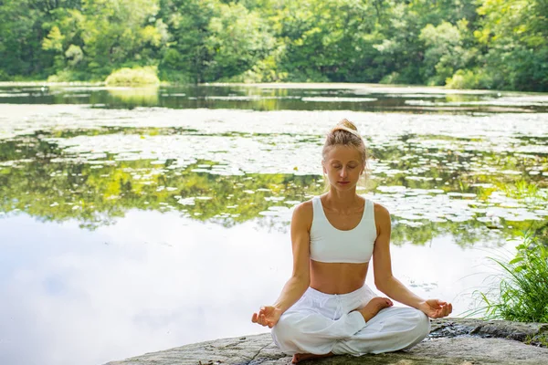 Mujer atractiva está practicando yoga sentado en pose de loto en piedra cerca del lago . — Foto de Stock