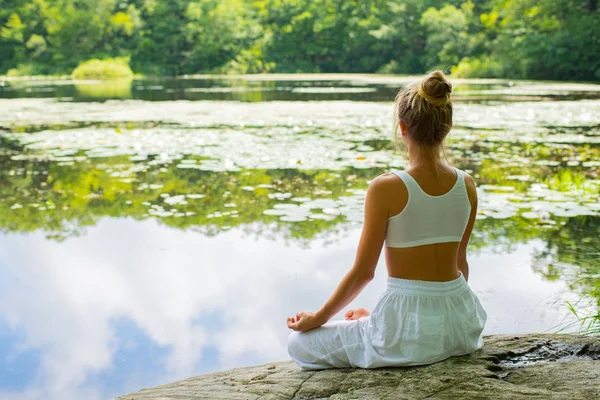 O femeie atrăgătoare practică yoga stând în lotus pozând pe piatră lângă lac . — Fotografie, imagine de stoc