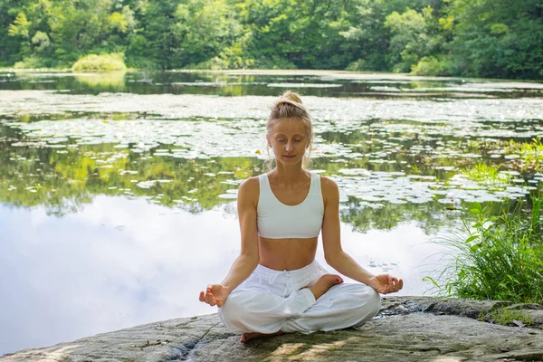 O femeie atrăgătoare practică yoga stând în lotus pozând pe piatră lângă lac . — Fotografie, imagine de stoc
