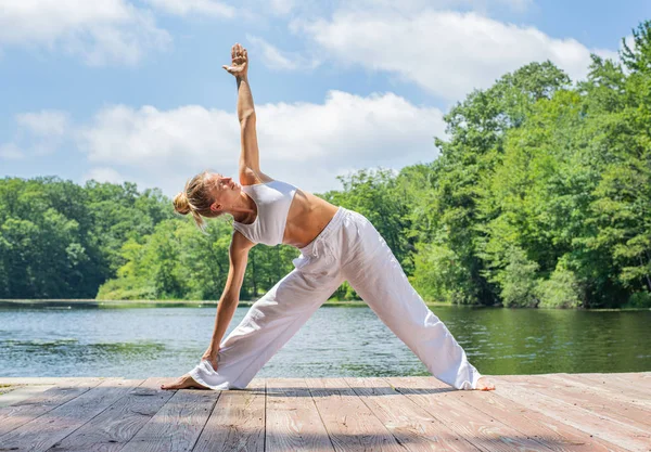 Jovem atraente está praticando ioga, fazendo Utthita Trikonasana pose perto do lago . — Fotografia de Stock