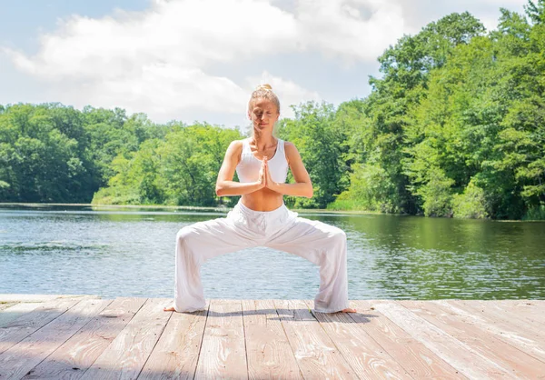 Femeia atractivă practică yoga, face exerciții Stupasana, stând în picioare în poziția Zeiței lângă lac . — Fotografie, imagine de stoc