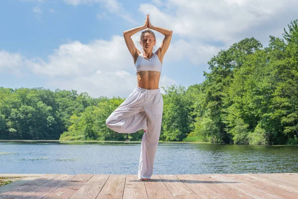 Femeia atractivă practică yoga, face exerciții fizice Vrksasana, stând în picioare în poză în copac lângă lac . — Fotografie, imagine de stoc