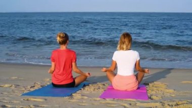 Genç kadınlar güneşin batımında sakin plajda yoga yapıyorlar. Meditasyon kızlar, Lotus oturan deniz kıyısında poz.