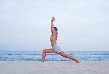 Sahilde yoga yapan genç bir kadın. Virabhadrasana Ben poz