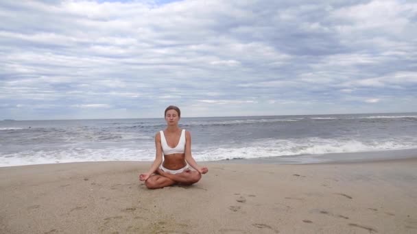 年轻女子正在海滩上练习瑜伽 动作慢动作 美丽的女孩做瑜伽在莲花姿势在海边 — 图库视频影像