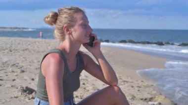 Kumsalda cep telefonuyla konuşan mutlu bir kadın. Doğanın tadını çıkaran kız sahilde tek başına oturuyor.,