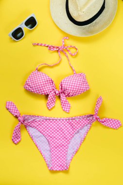 İki adet pembe damalı mayo, beyaz güneş gözlüğü ve hasır şapka sarı arka plan üzerinde üstten görünüm.