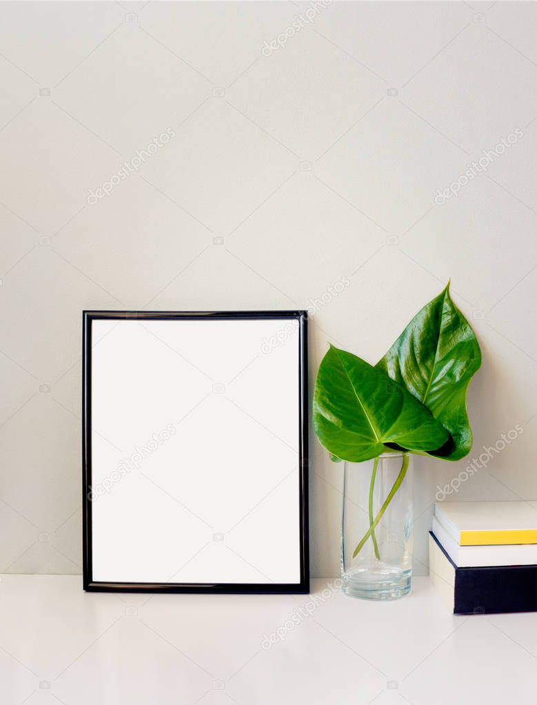 Black photo frame, green plant in a chrystal vase and a pile of books arranged against emty grey wall. Frame mock-up.