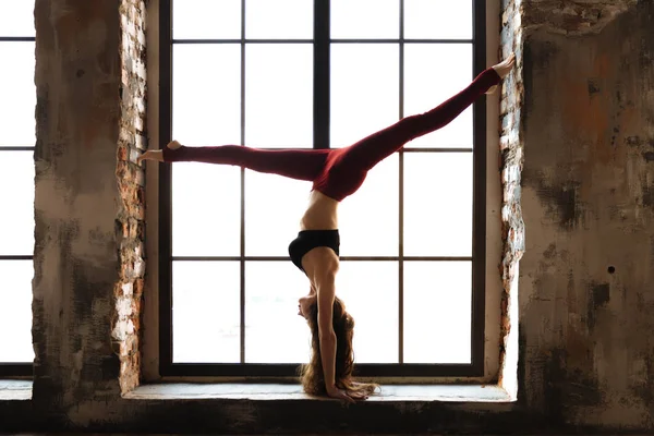 Yoga chica en una parte superior negra y rojo leggings está de pie en sus manos en la ventana de una ventana grande. — Foto de Stock