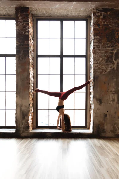 Menina Yoga em uma parte superior preta e leggings vermelhos está em suas mãos na janela de uma grande janela — Fotografia de Stock