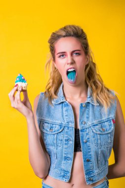 Deli kız dilini gösterir. Moda makyaj ve saç modeli. Yüz ifadesi, duygu, güzellik ve moda kavramı. Denim moda. Sarı izole kek ile seksi kız. Kız tadı cupcake