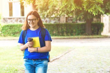 Güneşli bir günde Park'ta yürüyüş gözlüklü genç kız öğrenci. Hayattan zevk kahve kızla. Eğitim, yaşam tarzı ve insanlar kavramı. Okula dönüş. Defter tutan sırt çantası ile Gözlüklü kız