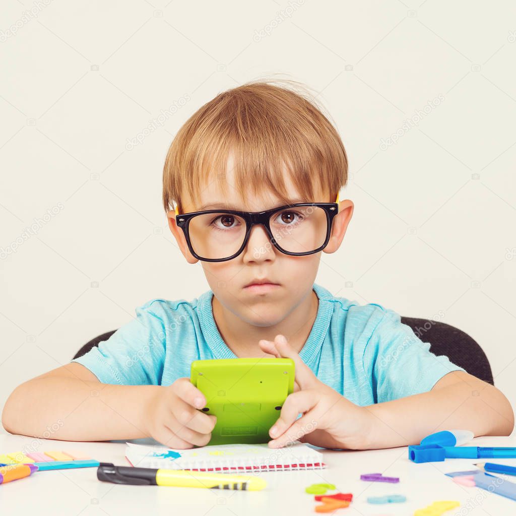Little business man with puzzled expression. Kid wearing big glasses and holding calculator. Kid sitting at the table with stationery. Smart kid. Little school boy in glasses doing homework